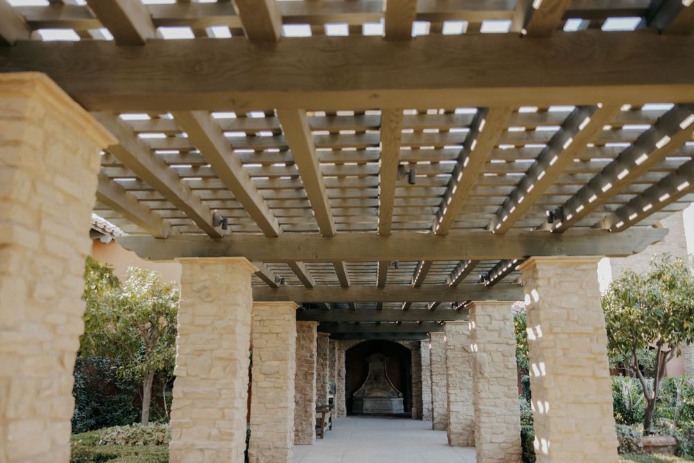Stone pillars and pergola create an elegant covered walkway entrance with natural lighting filtering through the slats.