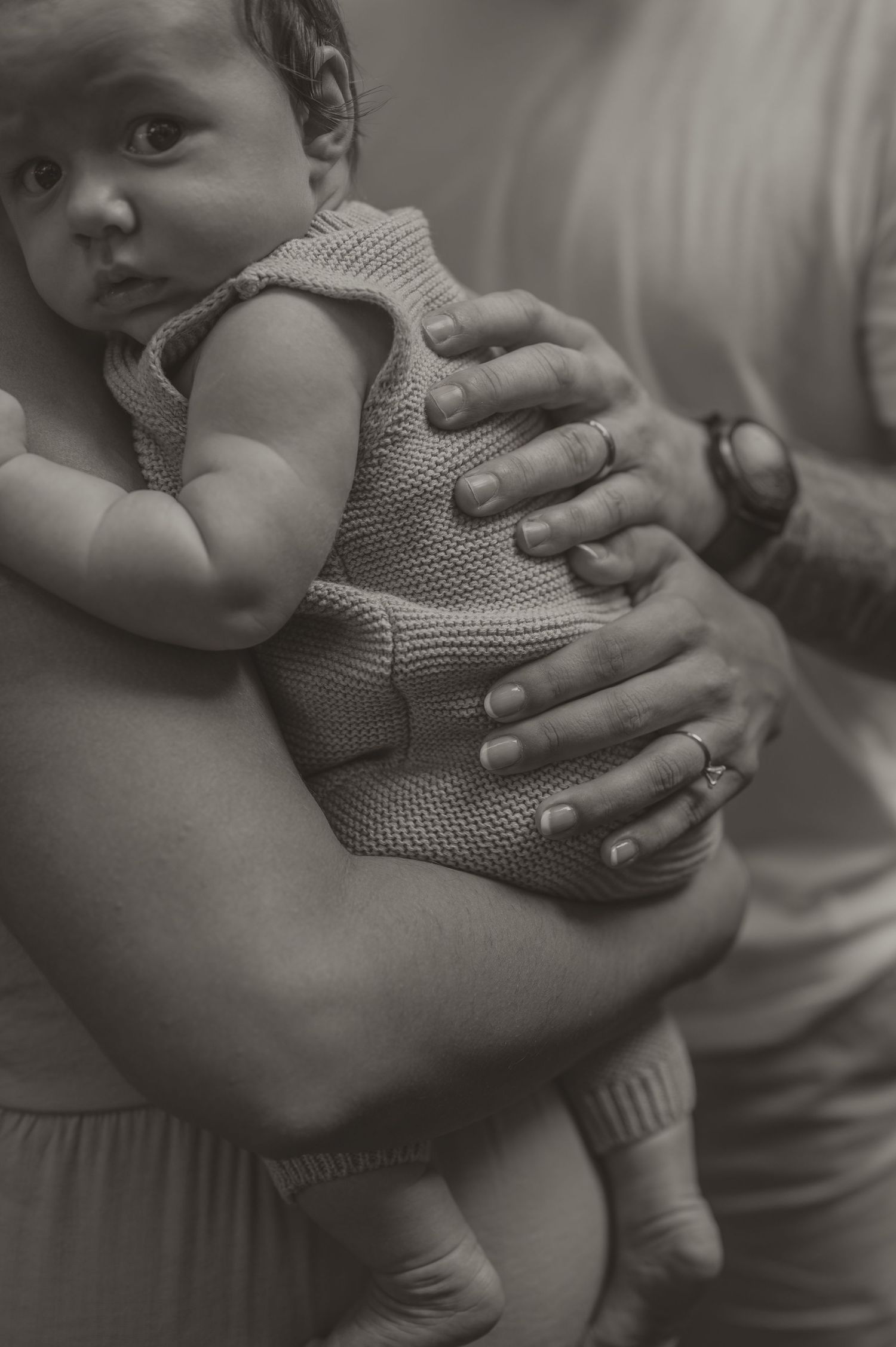 A black and white intimate photo by Samantha Campbell imagery showing gentle hands holding a baby wearing a knitted sweater.