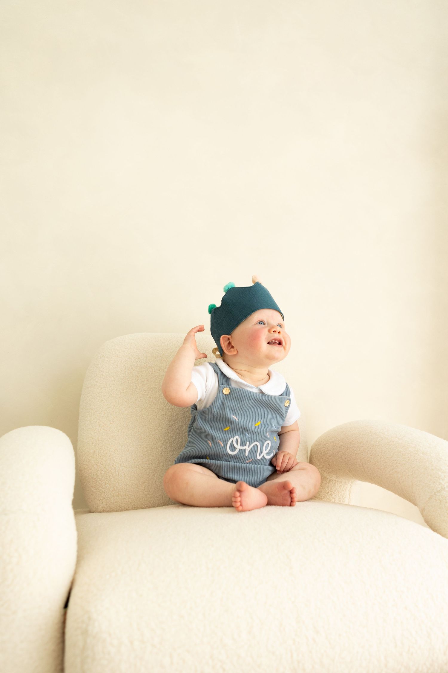 baby boy with his birthday outfit on smiling away sat on boucle chair with bright blue eyes.