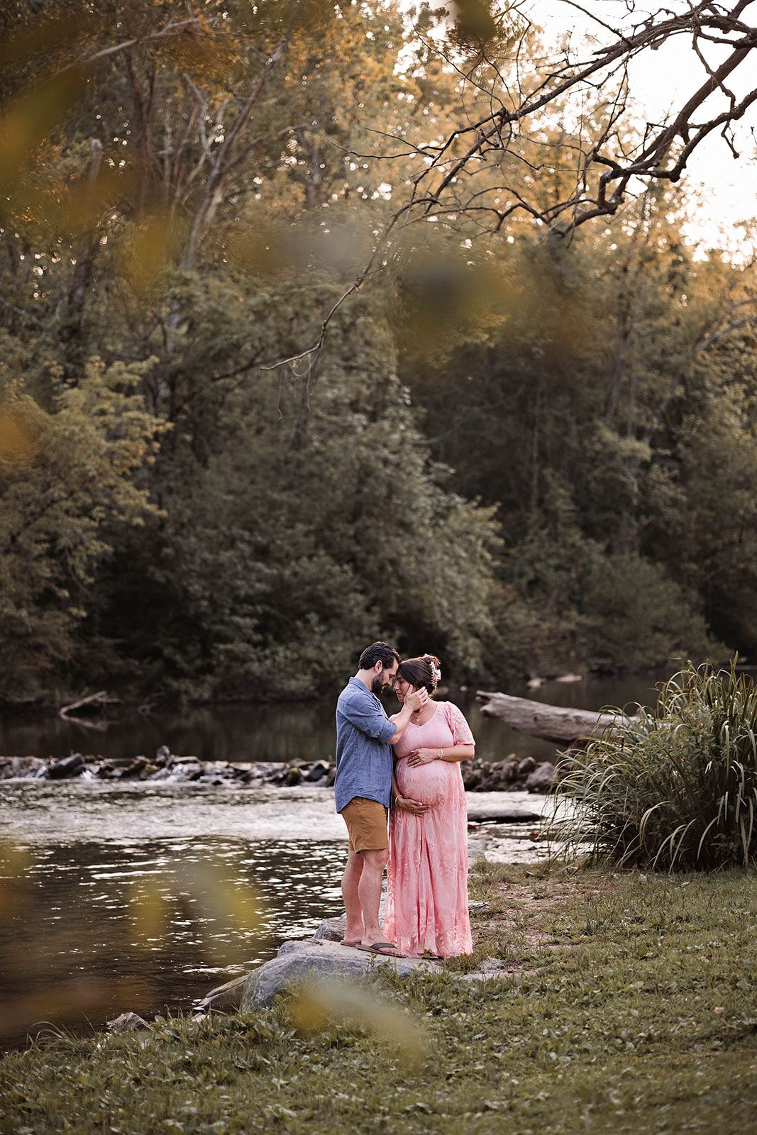 A pregnant couple shares a romantic moment by a tranquil river at sunset during a maternity photo session near Mechanicsburg PA.