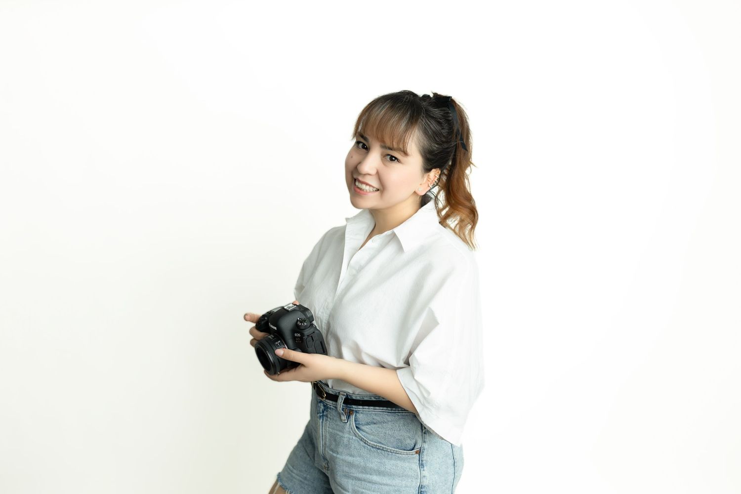Woman in white shirt and jeans holds black object while standing against white background.