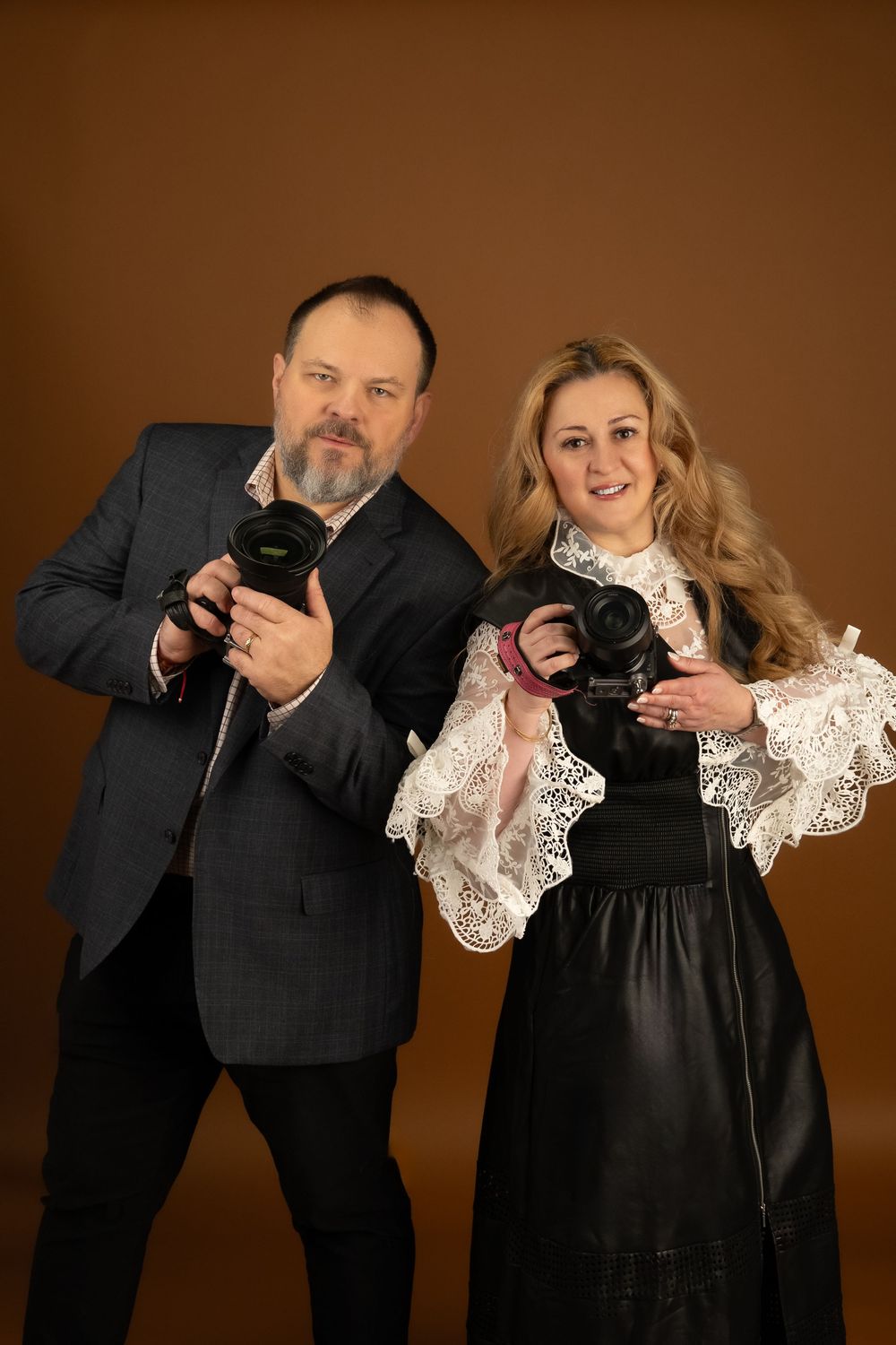 Professional photography duo posing with cameras against brown backdrop wearing formal attire.