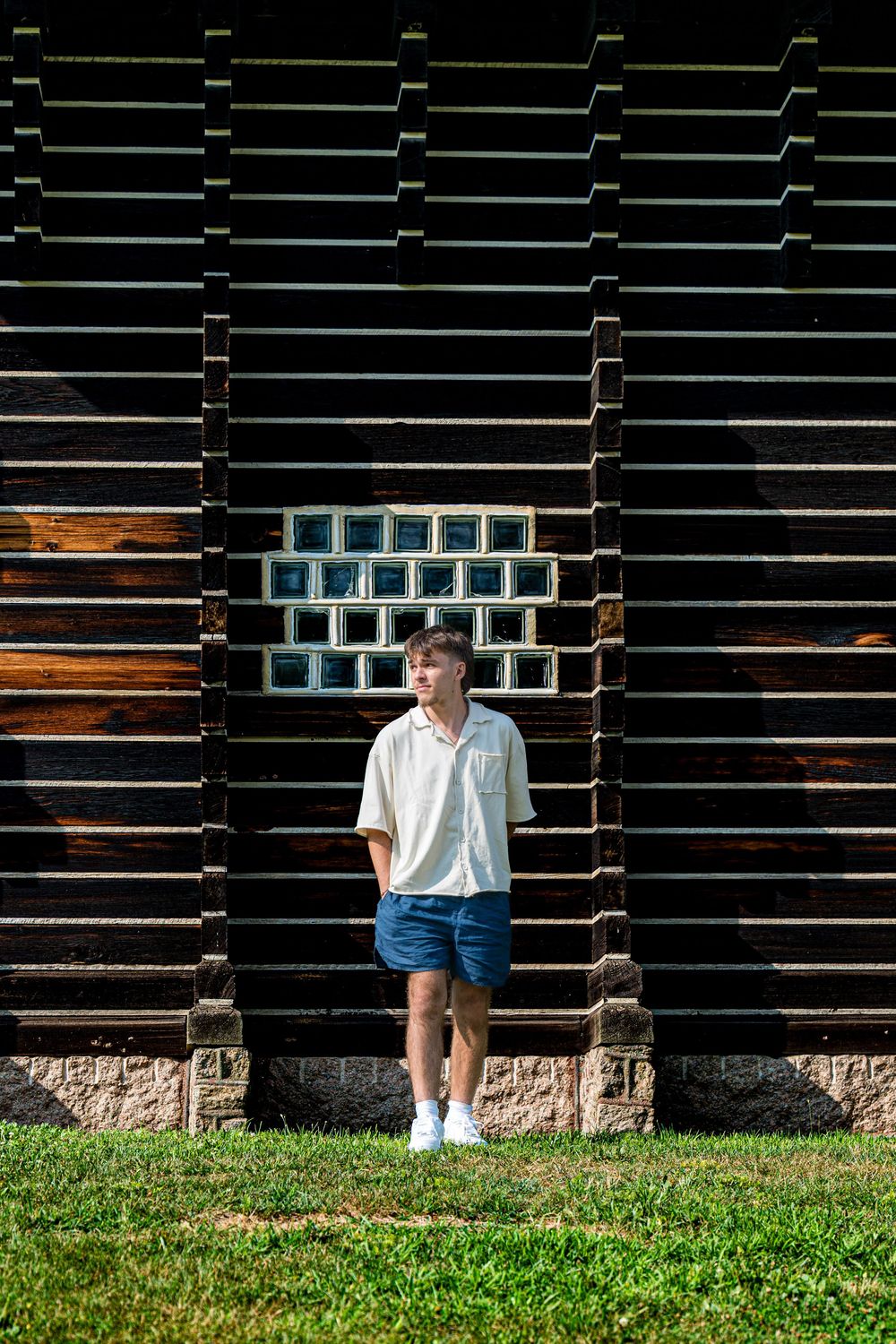 A person stands against a rustic dark wooden barn wall with grass in the foreground.
