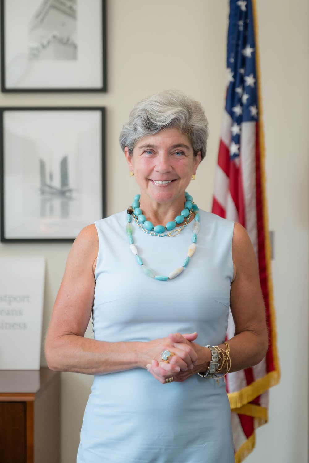 A woman in a light blue sleeveless top and turquoise necklace stands smiling in front of an American flag.
