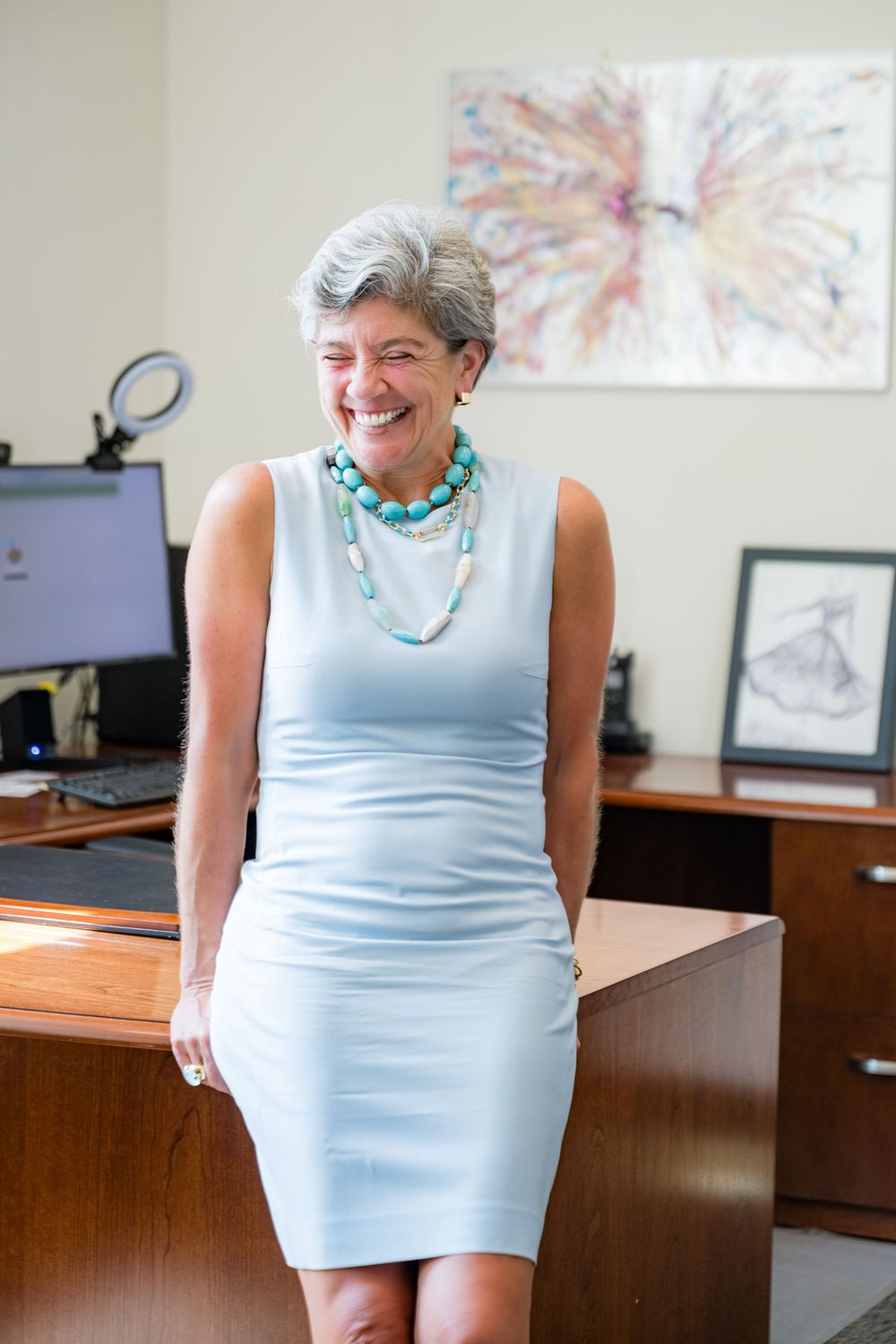 Professional in a light blue dress and turquoise beaded necklace stands in an office with artwork and computer visible.