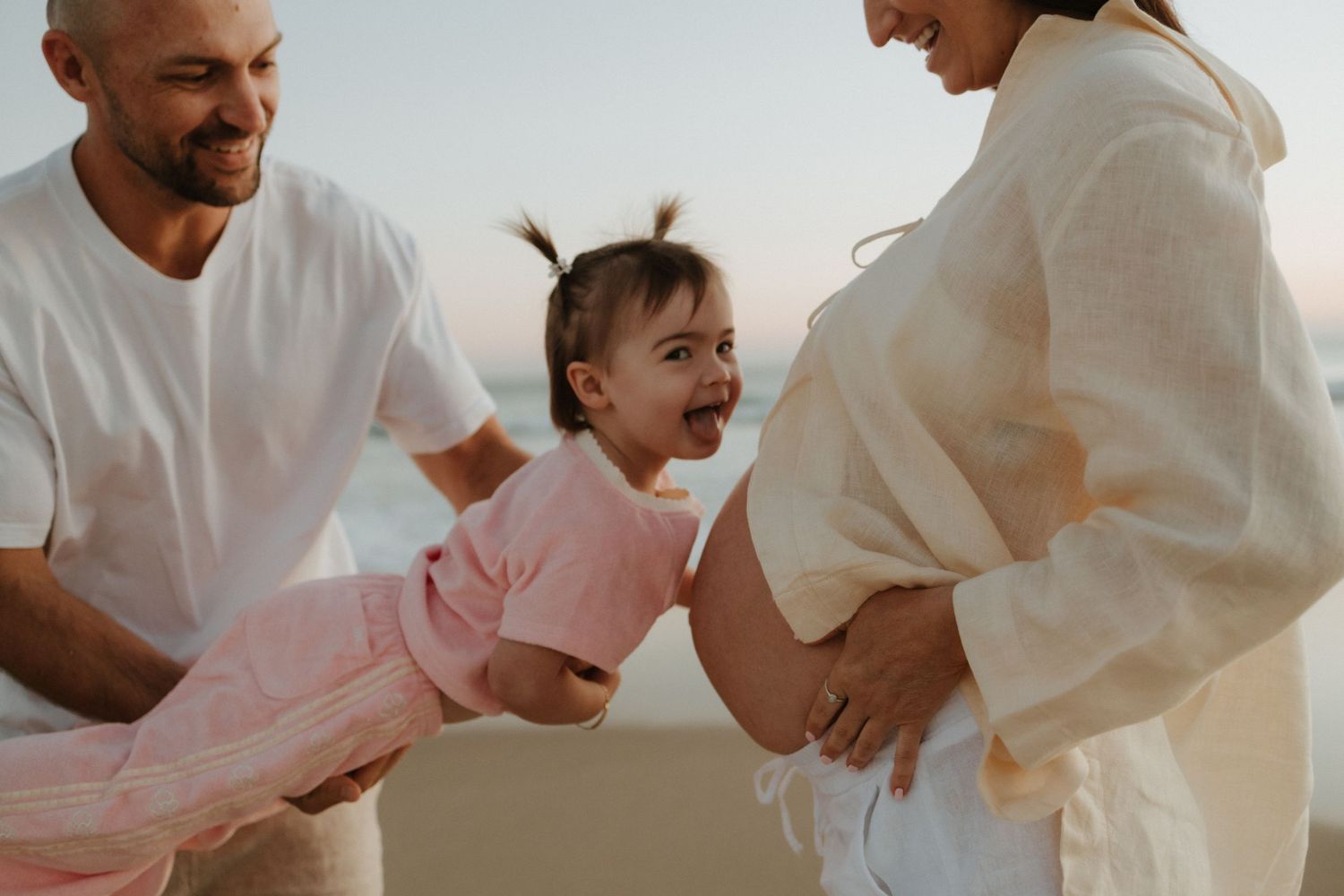 A joyful family moment captured in warm sunlight as parents playfully hold their laughing child wearing pink.