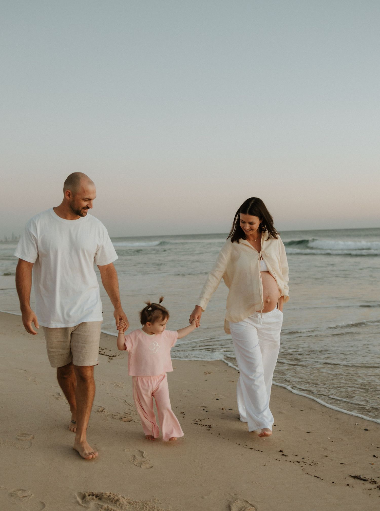 A family walks hand in hand along a beach at sunset with ocean waves in the background.