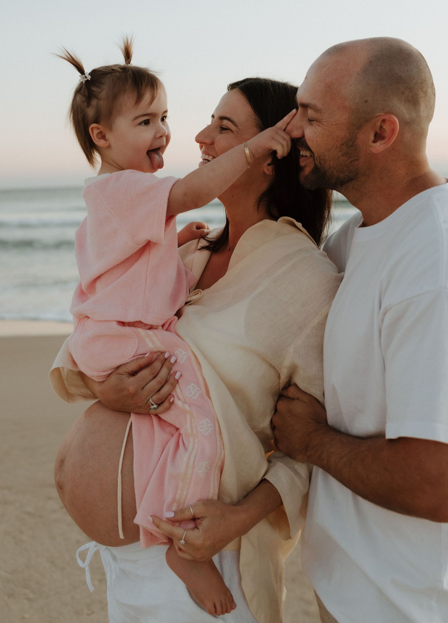 A loving family shares a tender moment together on the beach at sunset, with a pregnant mother in white.