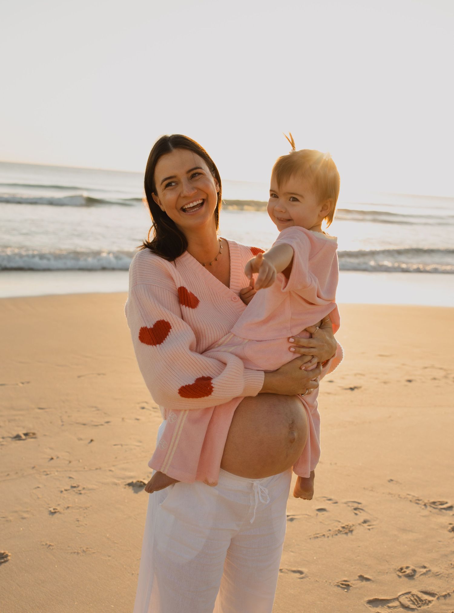 A parent and baby in matching white outfits share joyful moments together on a sunny beach.