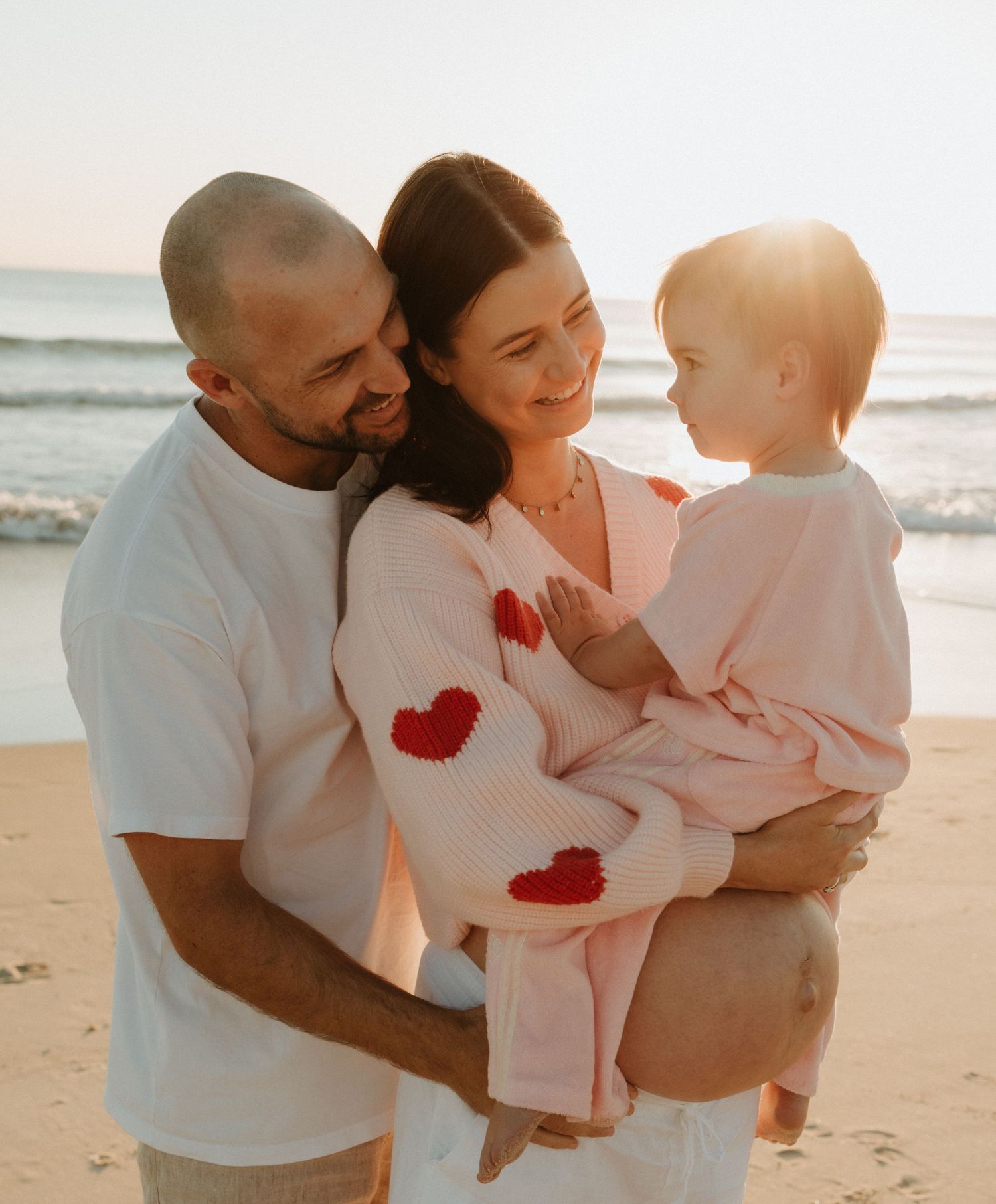 Family wearing matching white and red heart shirts embraces lovingly on a sunny beach at sunset.