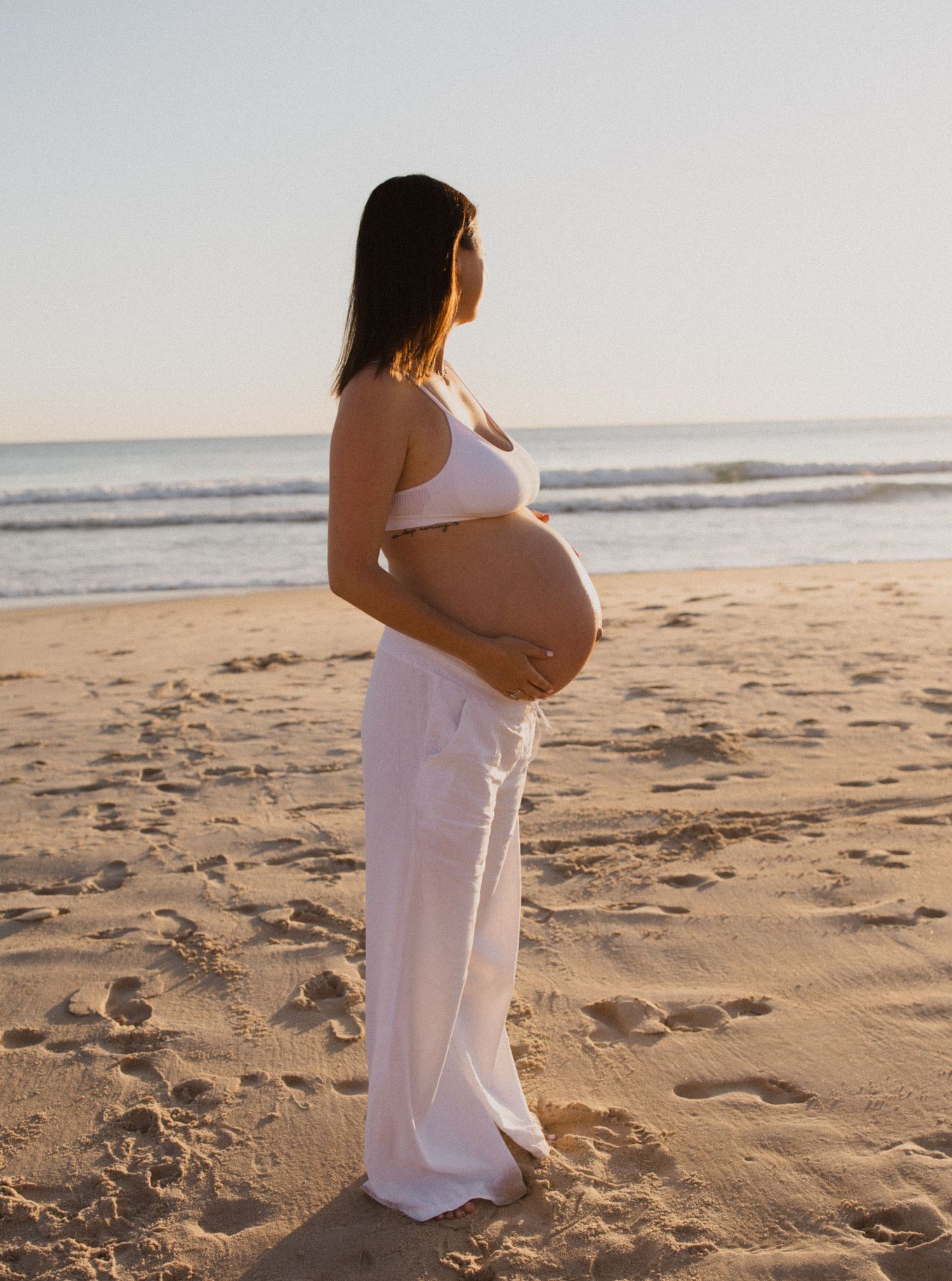 A person in a flowing white dress stands on a sandy beach at sunset, cradling their pregnant belly.