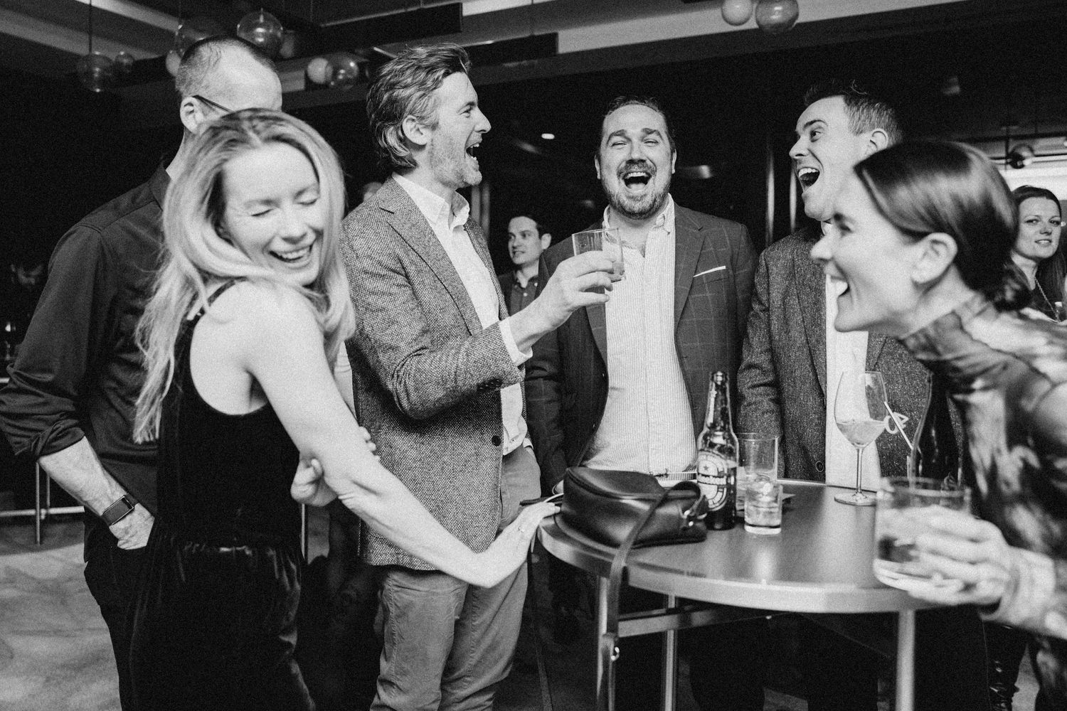 A joyful candid black and white photo of people laughing together at a social gathering in a bar setting.