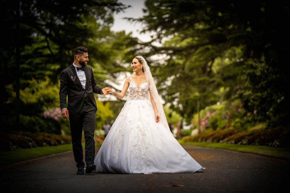 A wedding couple walks hand in hand down a tree-lined path in dramatic lighting.