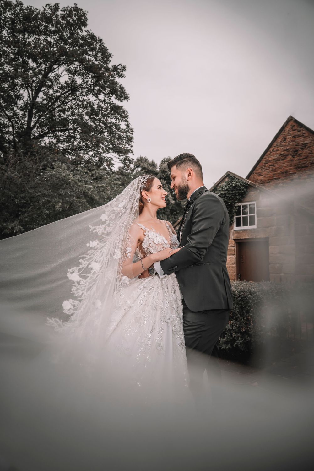 A romantic wedding portrait with flowing veil against a rustic brick building backdrop.
