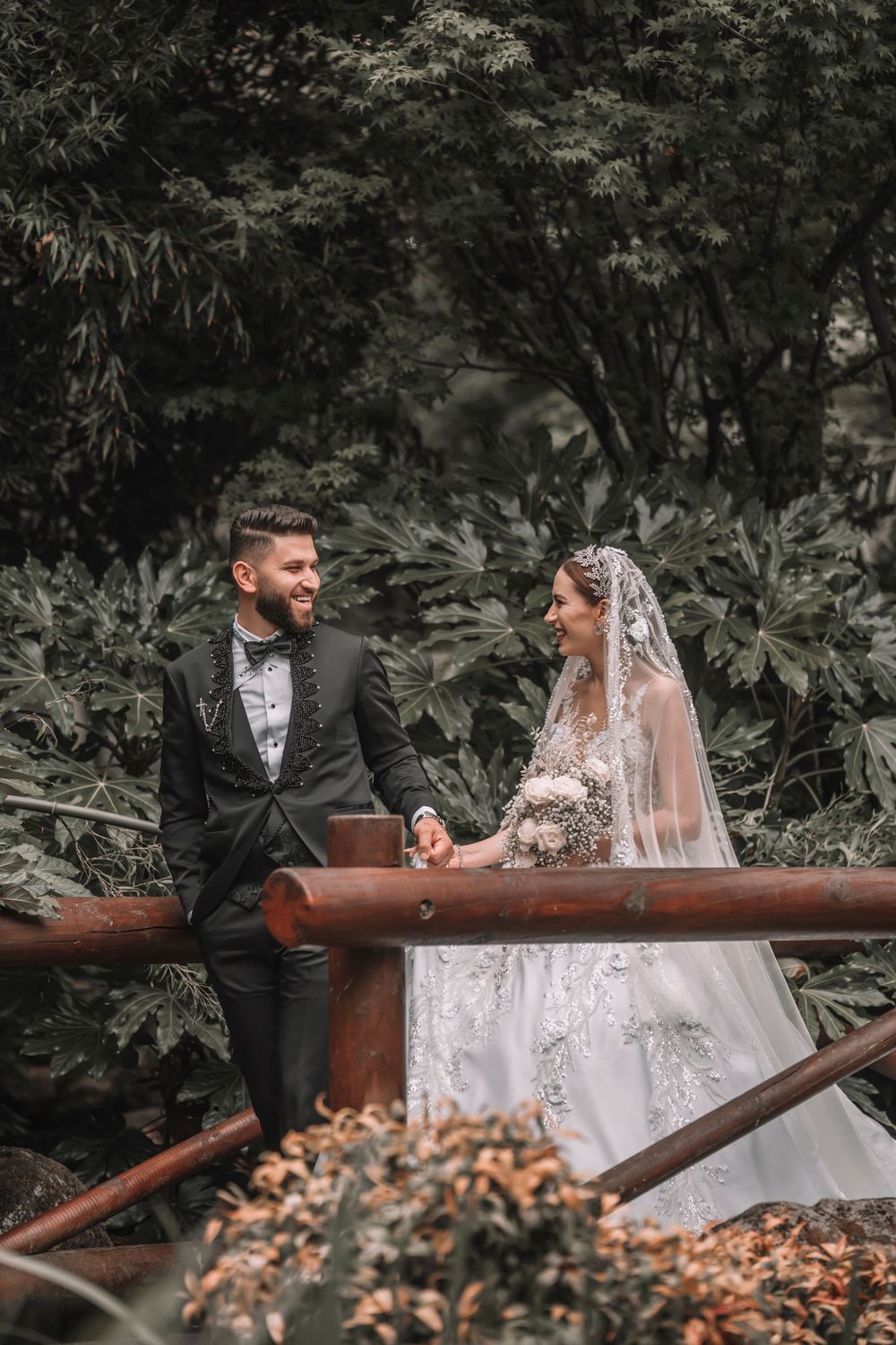 A newlywed couple shares a moment on a rustic wooden bridge surrounded by lush greenery.