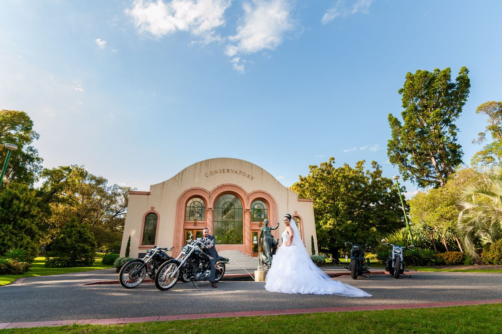 A bride in white poses with motorcycles outside a beautiful arched building on a sunny day.