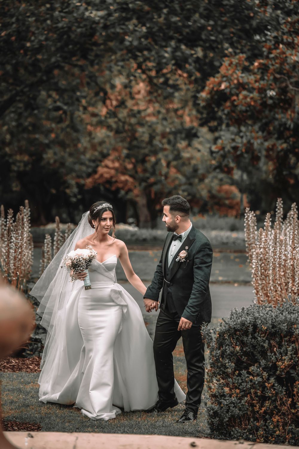 A wedding couple walks through an autumn garden with ornamental grasses and trees.