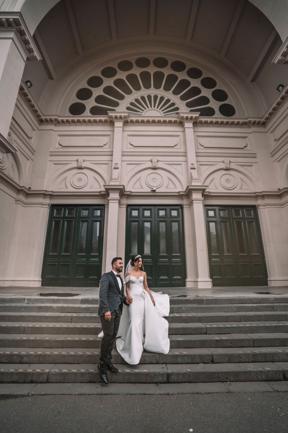 A wedding couple poses on steps of a historic building with ornate architectural details.