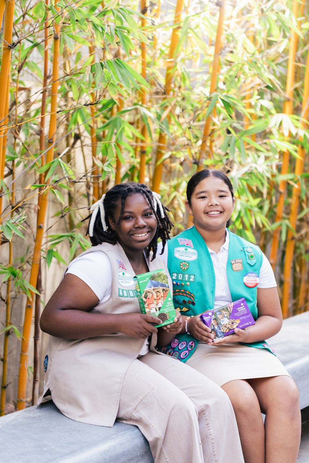 Two Girl Scouts wear uniforms and hold snacks while sitting together on a bench with bamboo plants in the background.