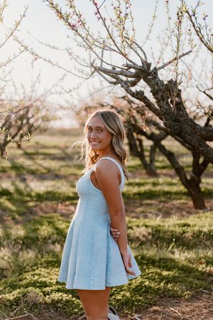 Woman in light blue sundress poses in orchard during springtime with blooming fruit trees at sunset.