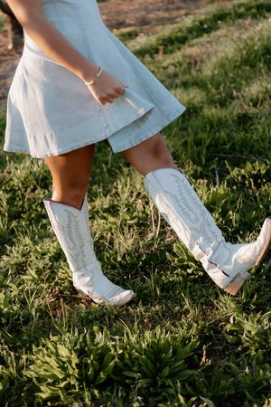 Series of photos showing white cowboy boots and flowing dress fabric walking through sunlit grass in a rustic outdoor setting.