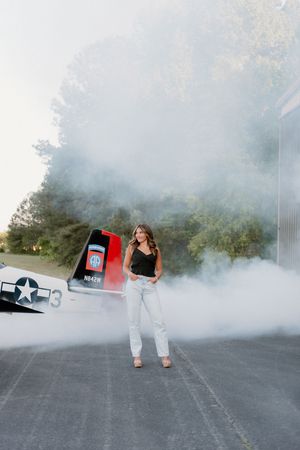 A person poses in a black tank top and light jeans amid smoke on a road with a vintage aircraft visible in background.