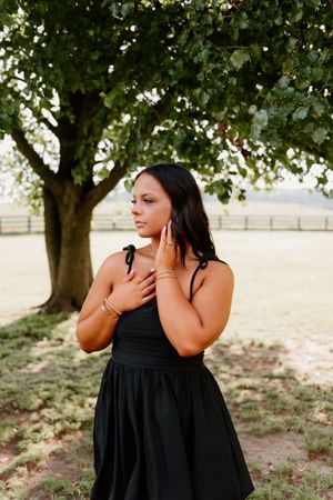 A person in a black dress poses gracefully under a large tree in an outdoor setting during golden hour.