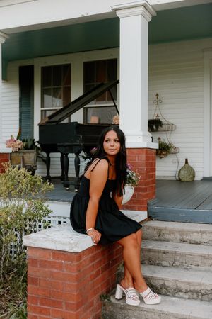 A person in a black dress sits on a brick porch step with a piano visible on the covered front porch.