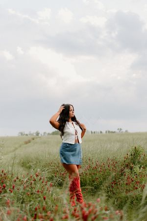 A person stands in a field of red wildflowers wearing a denim skirt and tall red boots under cloudy skies.