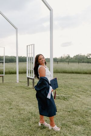 Someone poses in white top and navy cardigan on grassy field with white posts in background.