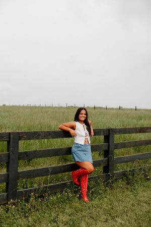 A person in red boots and denim skirt poses casually against a rustic wooden fence in a rural field.