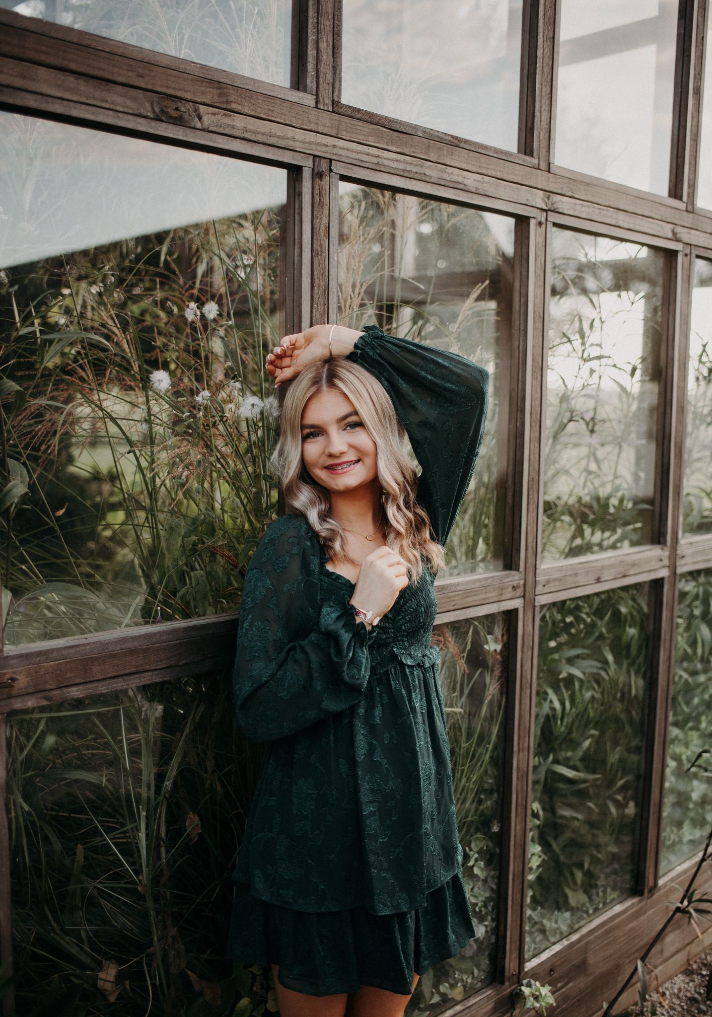 Young woman in emerald green dress poses against greenhouse windows surrounded by plants and natural light.