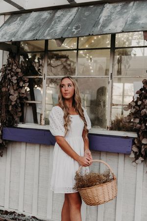 Person in white lace summer dress poses by rustic greenhouse with window boxes and woven basket.
