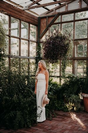 A sunlit conservatory interior with lush greenery and ferns surrounds someone in an elegant white dress holding a hat.