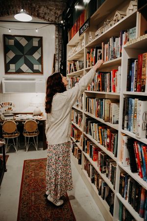 A person wearing a floral skirt reaches up to browse books on white shelves in a cozy library or bookstore.