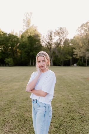 A person in a white t-shirt and light blue jeans poses thoughtfully in a grassy field at sunset.