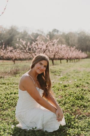A white sundress-clad woman sits in a field with blooming cherry blossom trees in the background at sunset.