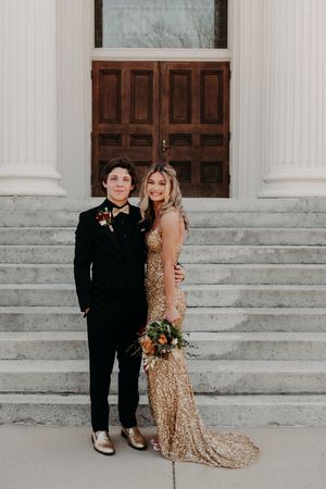 Two people in formal attire pose together on stone steps in front of a wooden door at a grand building entrance.