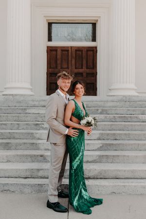 Elegant couple poses on marble steps, with one wearing an emerald sequin gown and the other in a light grey suit.