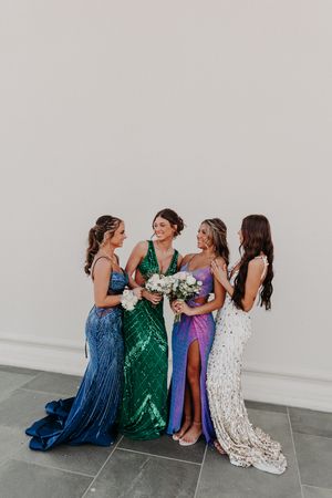 Four women in elegant sequined gowns pose together holding bouquets against a white wall at a formal event.