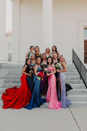A celebratory group wearing formal evening gowns in red, blue, and pink poses together on white stone steps.