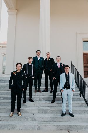 A group in formal black attire poses casually on white stone steps in front of a building with columns.