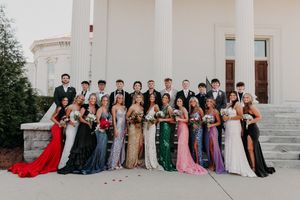 A group dressed in formal attire, dresses and gowns, sitting in formation on the steps of a grand building.