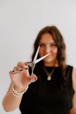 Professional hairstylist in black top with pearl bracelet holds up silver scissors against white background.