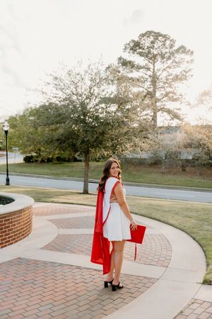 A figure in a white dress and red cape walks along a curved brick pathway through a green campus landscape at sunset.