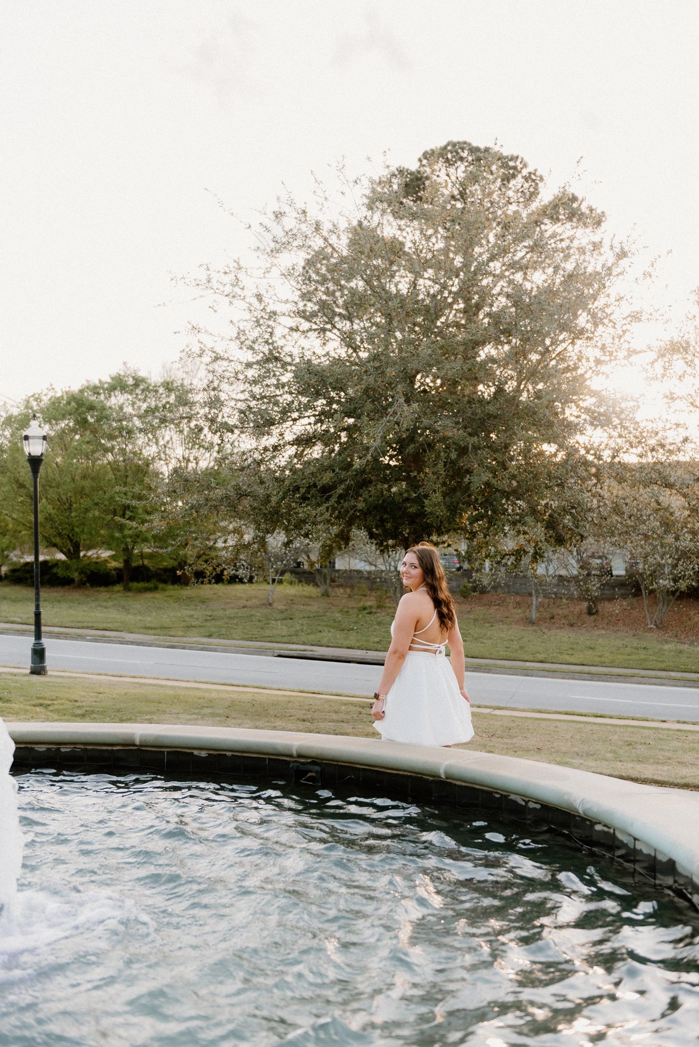 A person in a white dress sits at the edge of a fountain pool in a park setting during golden hour.