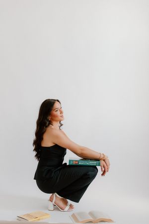 Person in black dress squatting down with books scattered around them on a minimalist white backdrop.