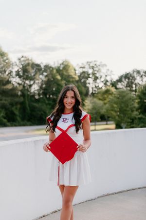 A graduate celebrates their academic achievement in a white dress with a red diploma holder outdoors on a sunny day.