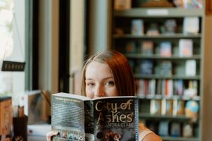 Person with red hair reading a City of Ashes book in front of a blurred bookshelf background.