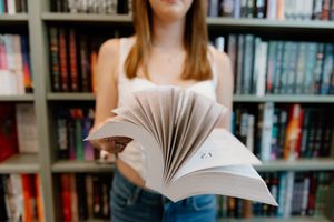 Someone flips through pages of an open book in a library with filled bookshelves in the background.